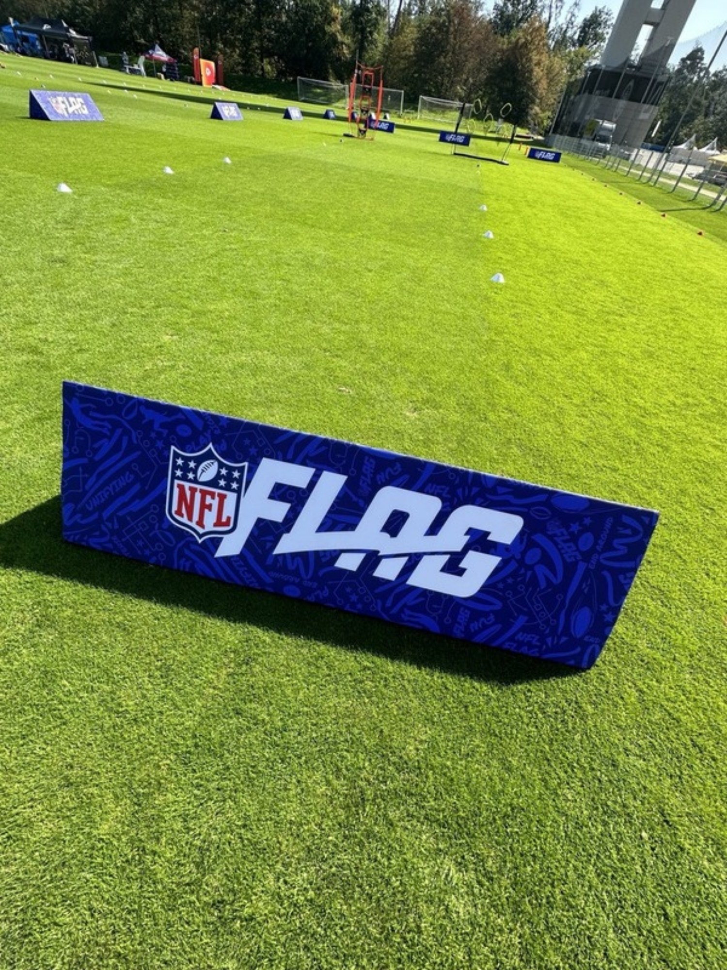 Blauer NFL-FLAG-Aufsteller auf einem Rasenplatz, im Hintergrund weitere Aufsteller und Hütchen auf dem Trainingsfeld