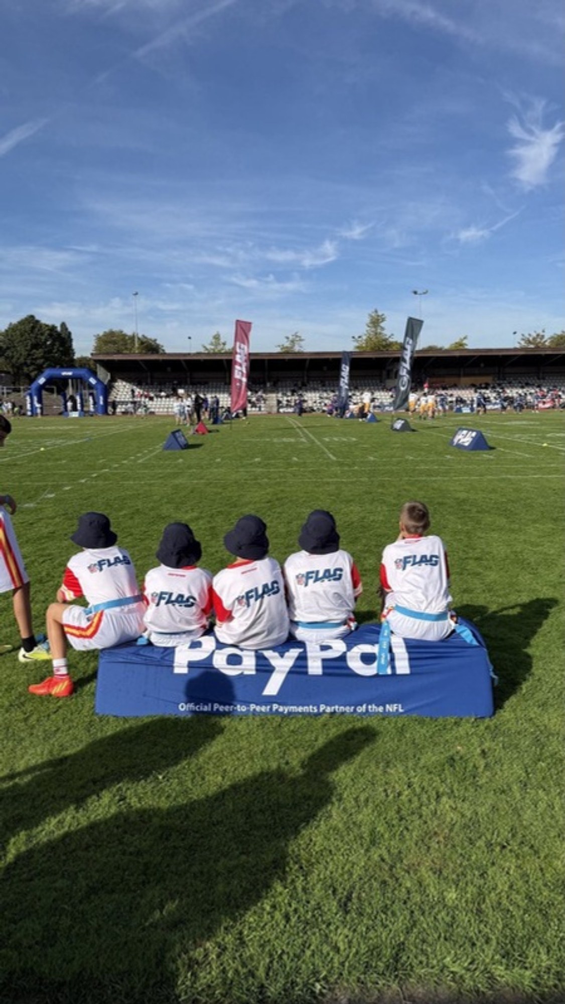 Fünf Kinder in NFL-FLAG-Trikots sitzen von hinten gesehen auf einer Bank am Spielfeldrand, im Hintergrund läuft ein Turnier