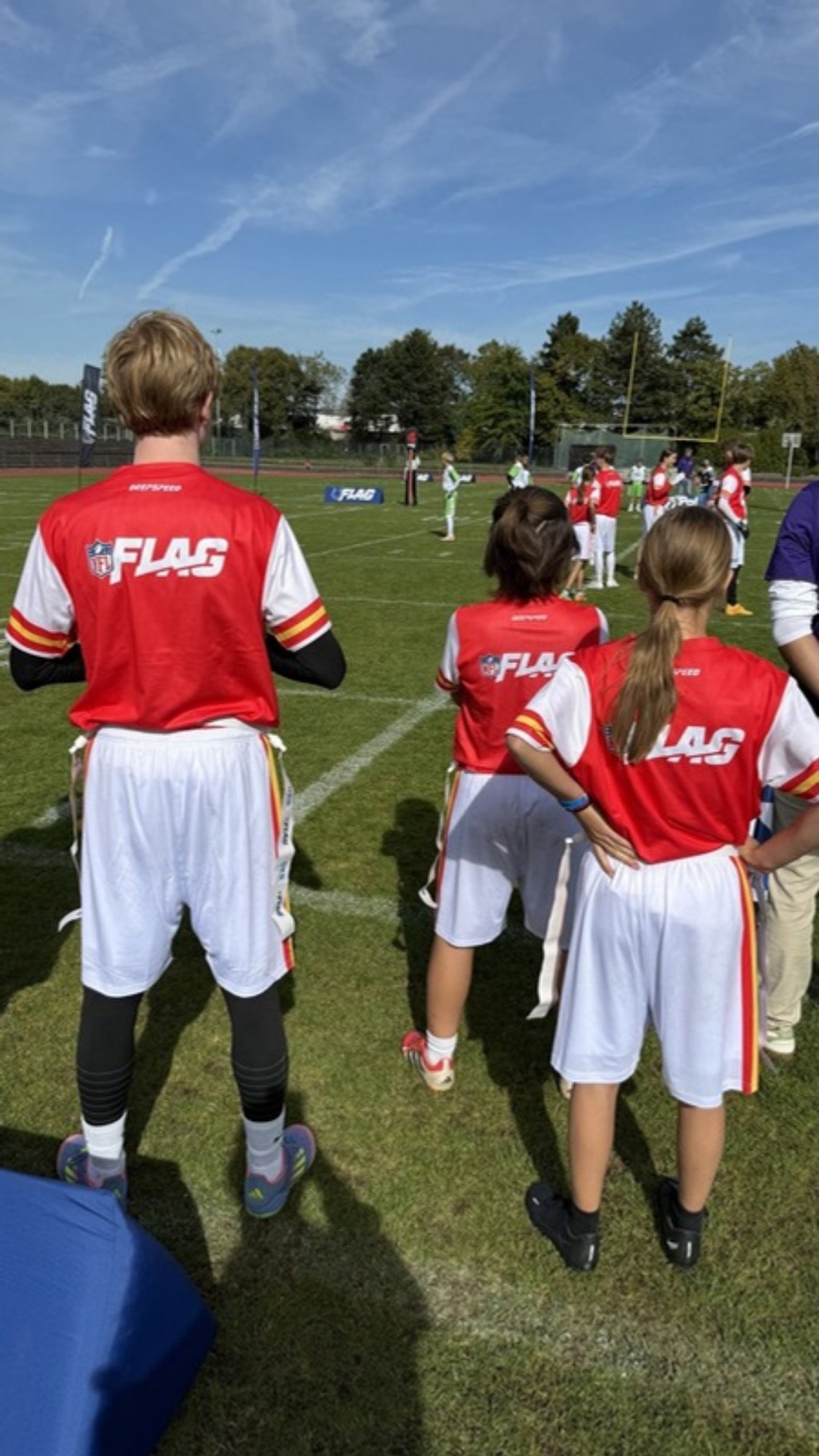Kinder und Jugendliche in rot-weißen NFL-FLAG-Trikots stehen von hinten gesehen am Spielfeldrand eines NFL-Flag-Turniers